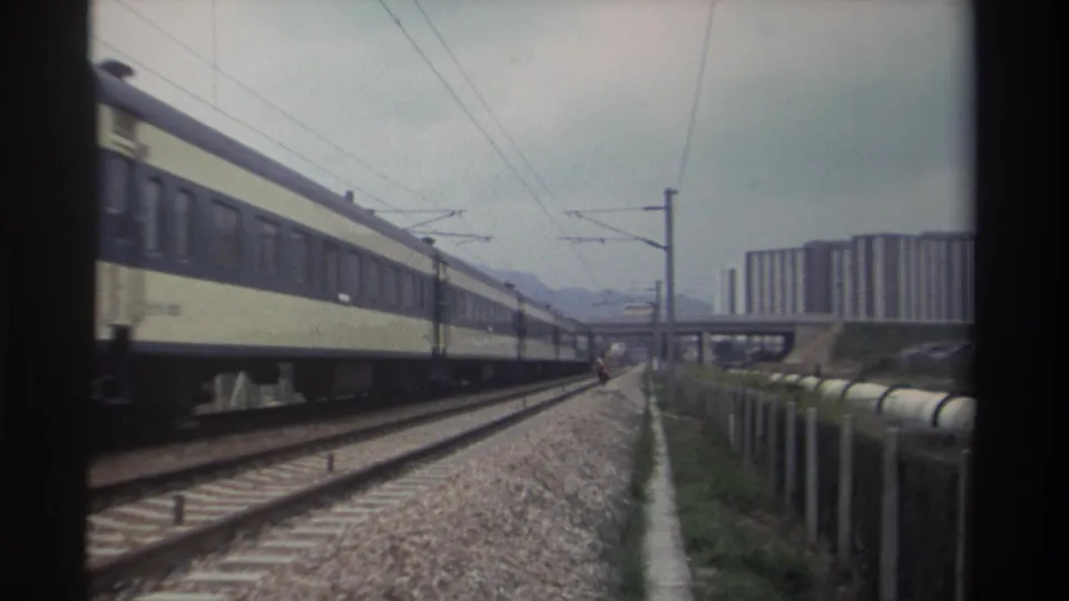 Cream and blue Taiwanese passenger train on electrified tracks with catenary poles, high-rise apartments, and distant hills, circa 1982.