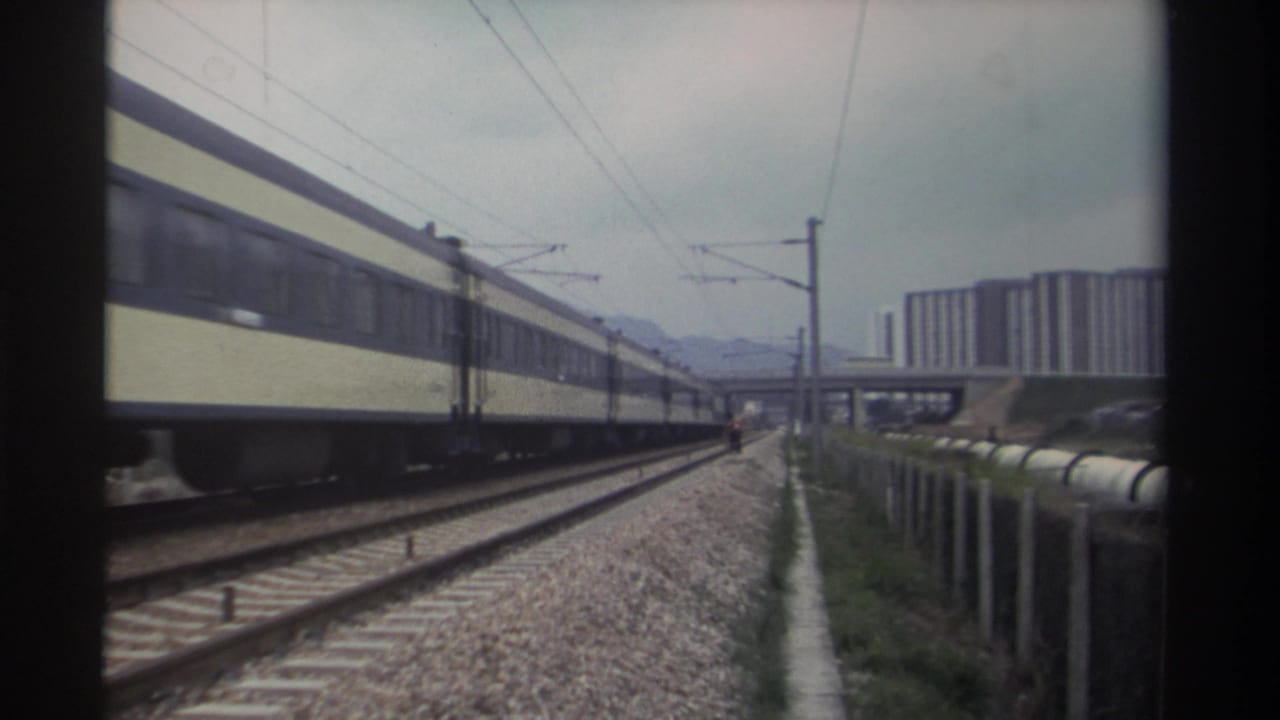 Long passenger train in cream and blue livery traveling along electrified tracks, apartment towers visible in background.
