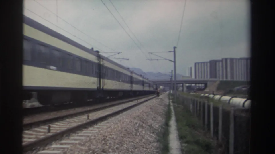 Long cream-and-blue passenger train on electrified rail corridor in Taiwan circa 1982, with concrete residential towers and mountains in background.