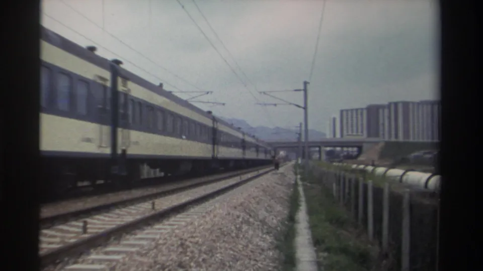 Tail end of a Taiwanese passenger train clearing frame circa 1982; open tracks, catenary poles, overpass bridge, and residential towers remain in view.