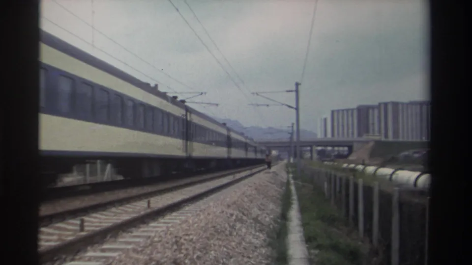 Cream and blue electrified passenger train departing at frame right in Taiwan circa 1982; overhead wires, open tracks, and residential high-rises remain in view.