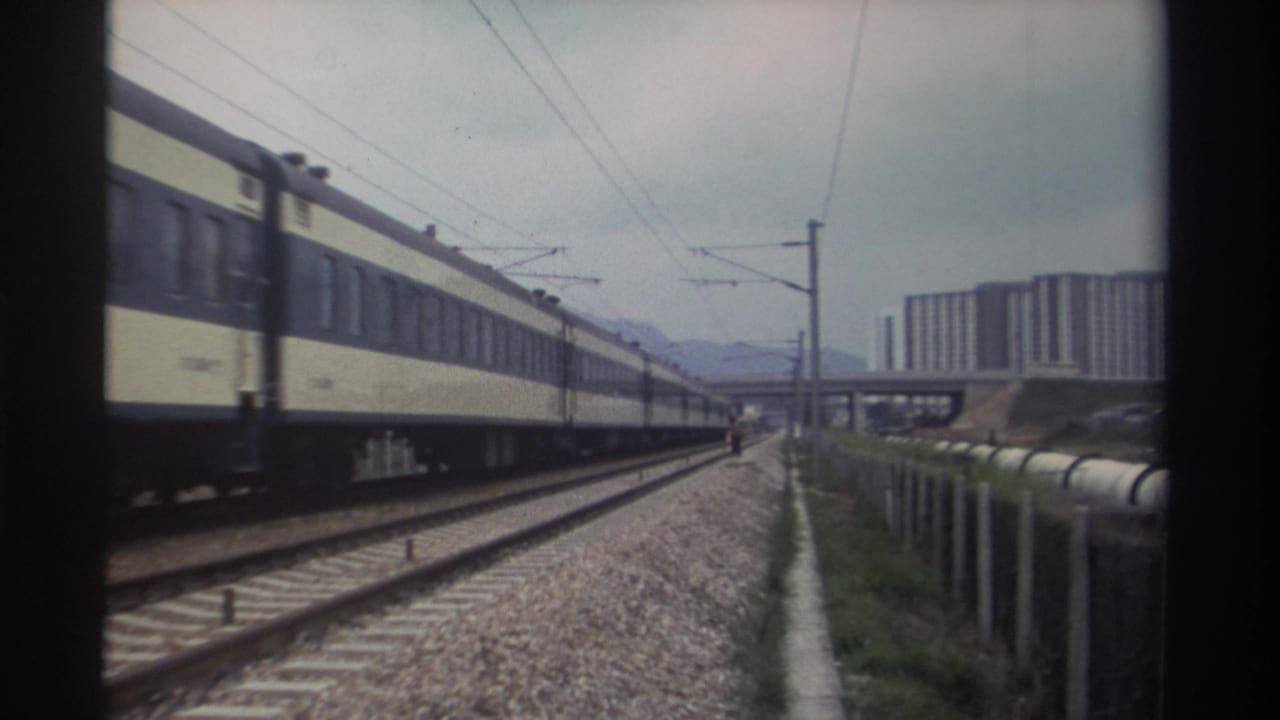 Cream and dark-blue passenger train passing along electrified tracks with high-rise buildings and mountains in background.