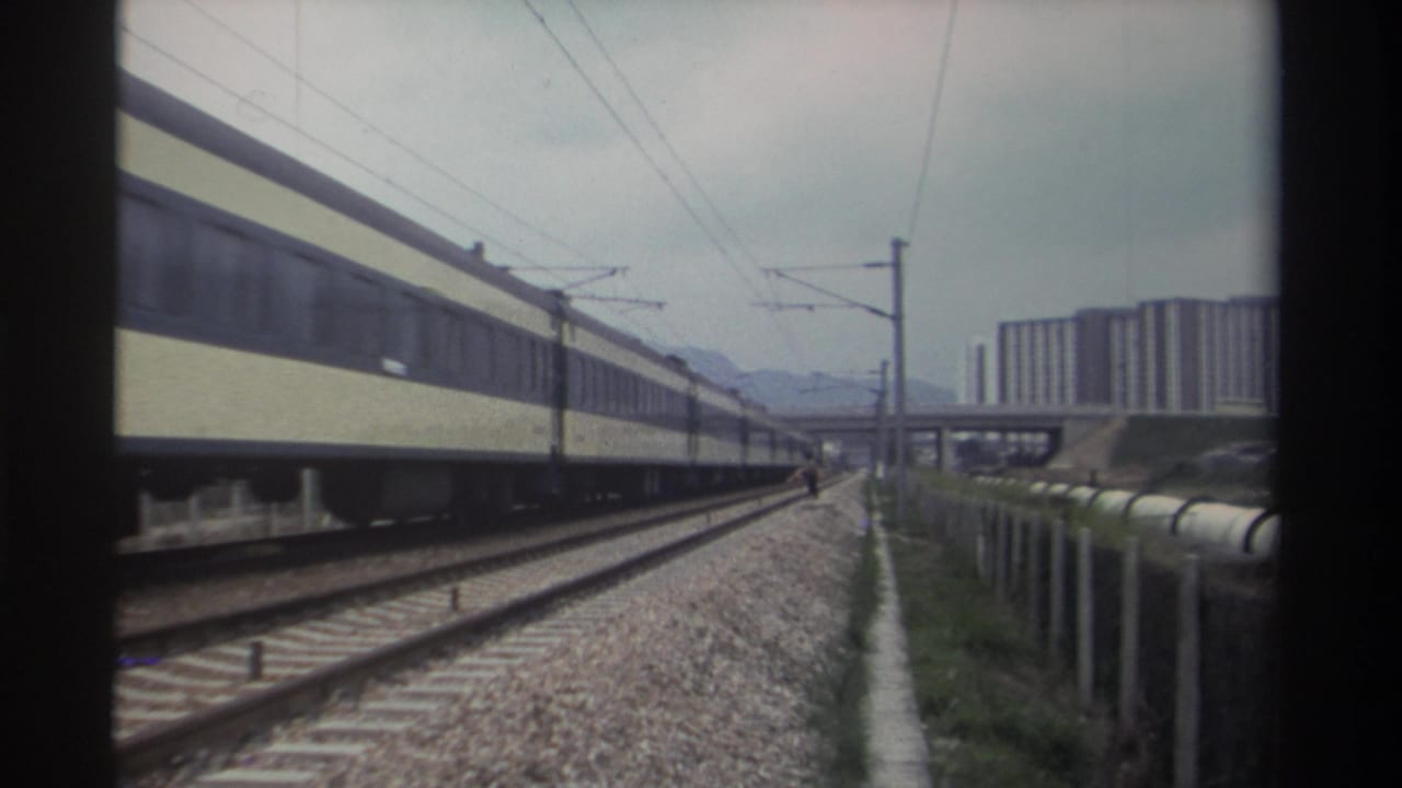 8mm film frame from Taiwan, circa 1982: passenger train with cream and dark-blue livery on electrified tracks, modernist residential towers and hazy mountains beyond.
