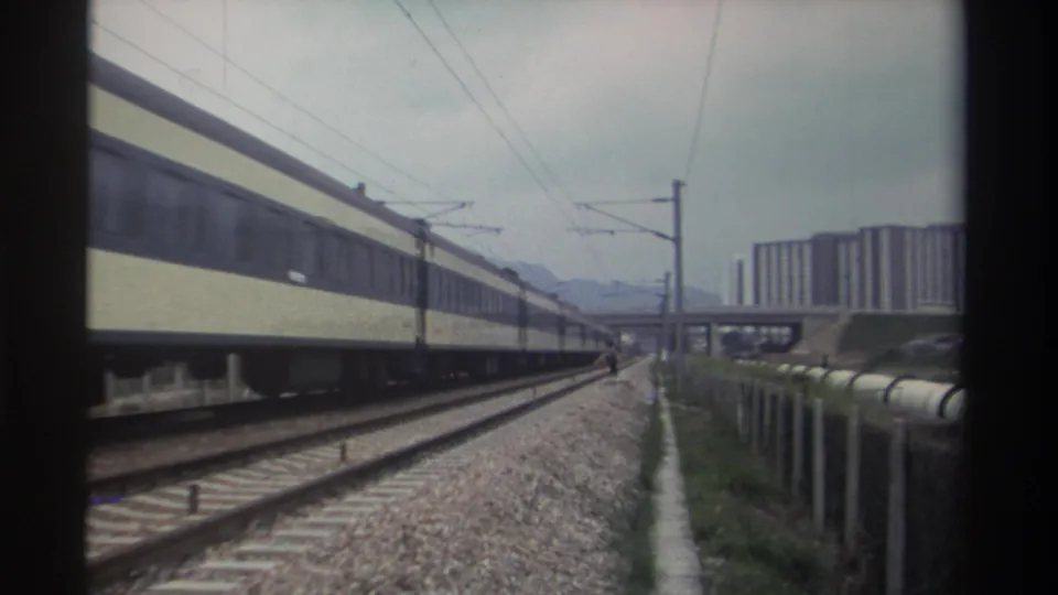 8mm film frame from Taiwan, circa 1982: passenger train with cream and dark-blue livery on electrified tracks, modernist residential towers and hazy mountains beyond.