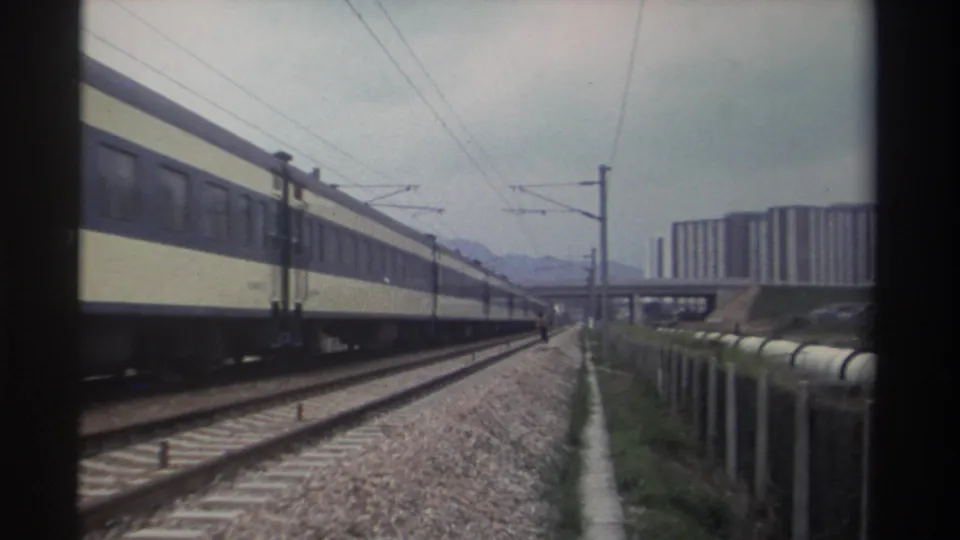 8mm home movie frame from Taiwan, circa 1982: cream-and-navy passenger carriages passing along electrified multi-track line, residential towers and mountain silhouette beyond.