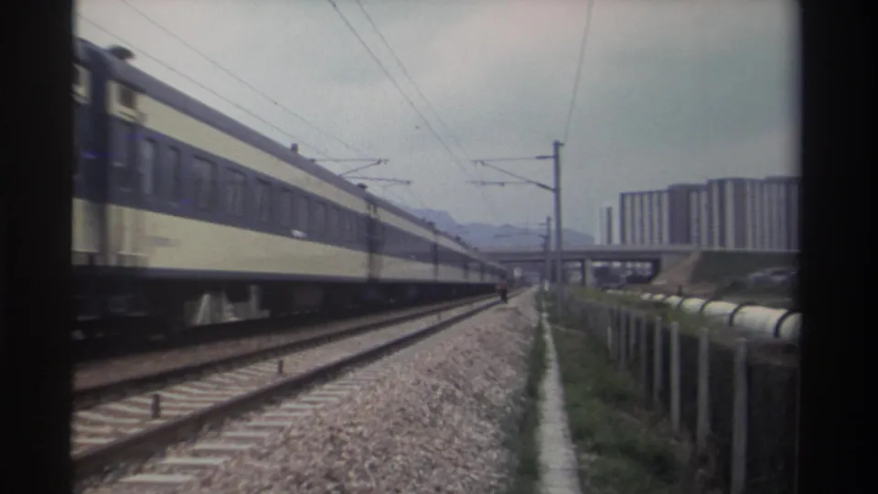Final 8mm frame from Taiwan, circa 1982: cream-and-navy passenger carriages on electrified tracks, blocky high-rise towers and hazy mountain range in the distance.