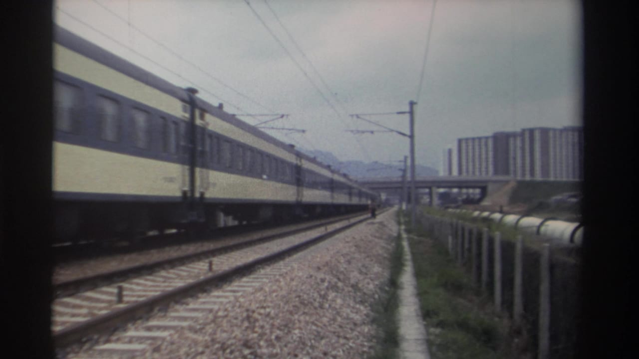Yellow and cream passenger train passing along tracks, concrete overpass and high-rise apartment blocks in background, overcast sky.