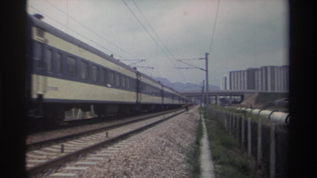 Yellow and cream passenger train moving away along electrified tracks in Taiwan, circa 1982; urban housing blocks and a concrete bridge visible in background.