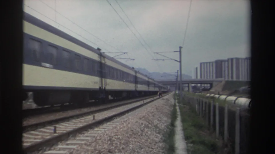 Passenger train receding further along electrified railway in Taiwan, circa 1982; gravel ballast, catenary poles, and high-rise housing blocks fill the frame.