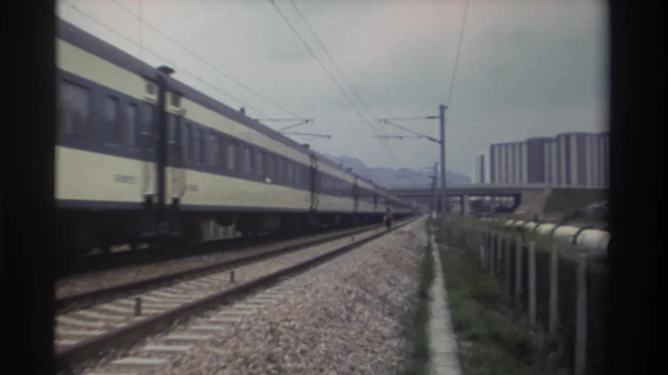 Electrified railway tracks in Taiwan, circa 1982, with a receding passenger train, high-rise housing blocks, and hazy hills visible under an overcast sky.