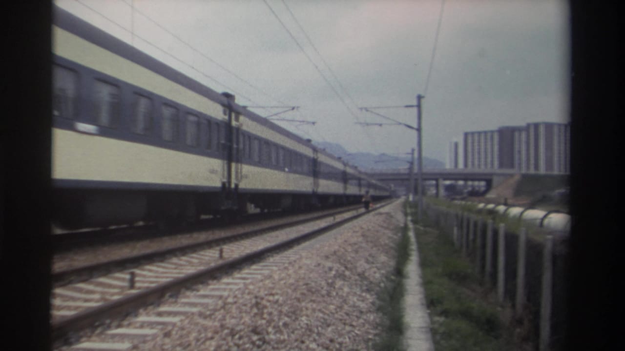 Cream and dark blue passenger coaches on electrified railway, circa 1982 Taiwan; apartment blocks and distant mountains visible in background.