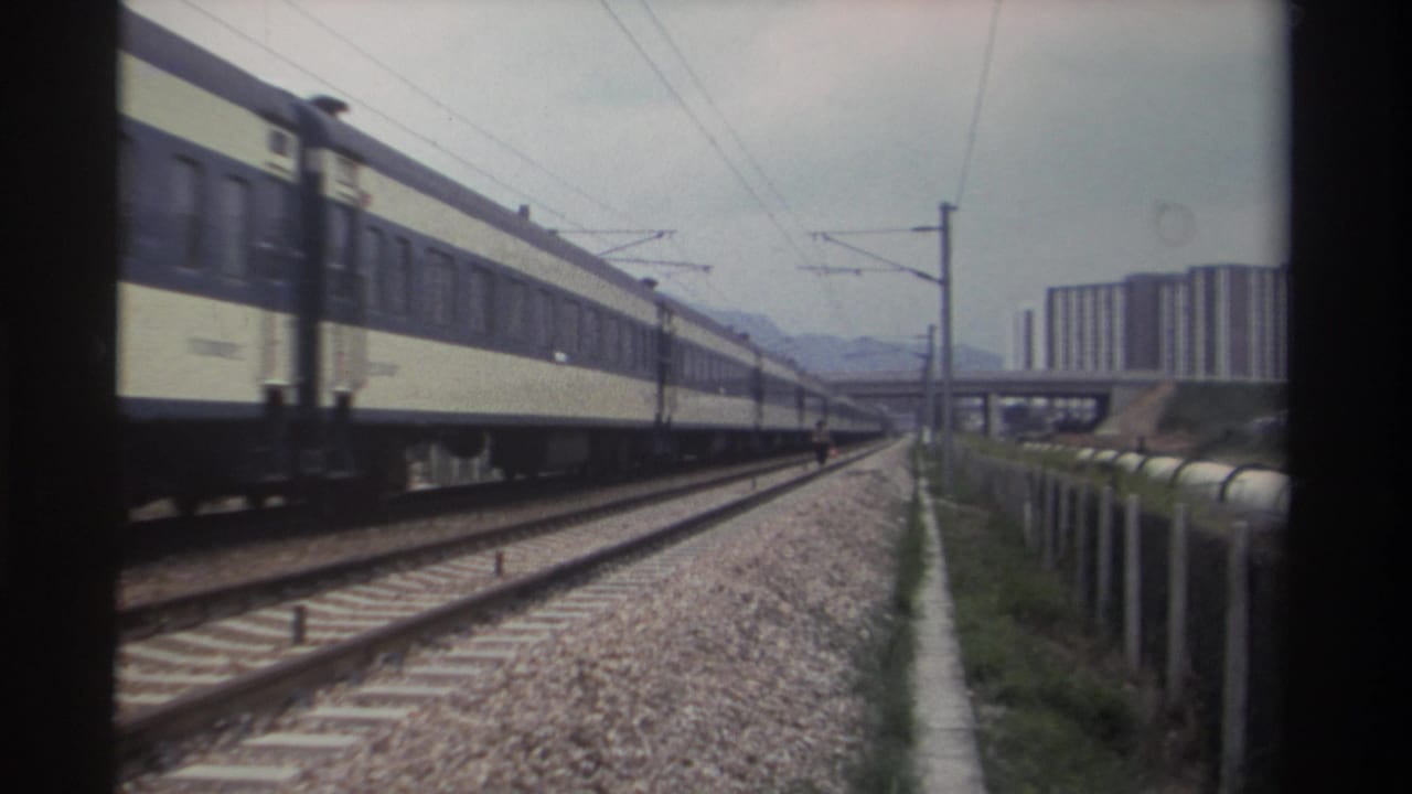Passenger train with cream upper and dark blue lower livery on electrified double track; apartment towers and hazy mountains in distance.