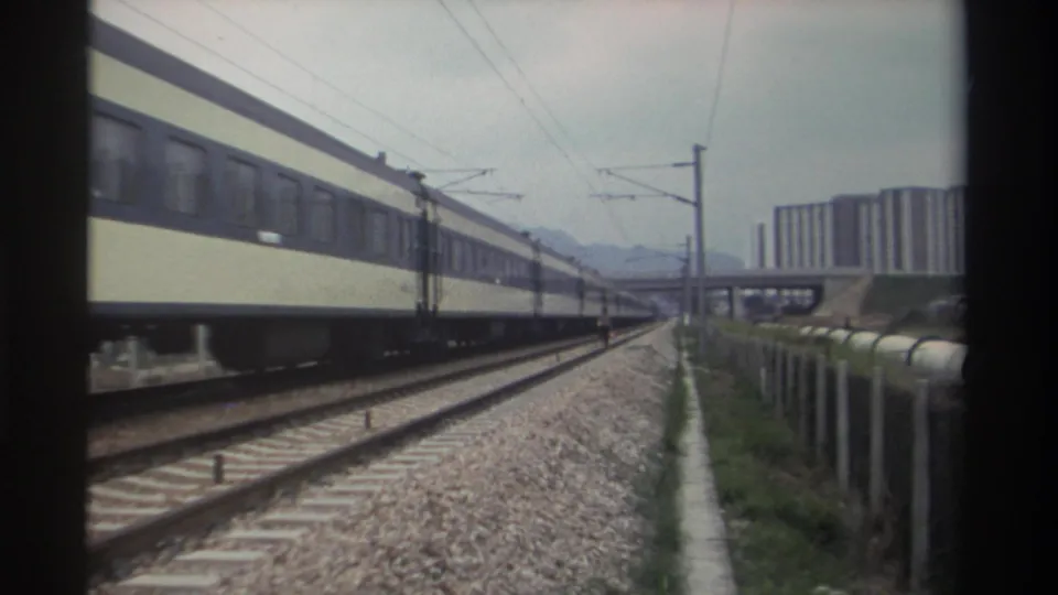 Cream-and-blue passenger coaches passing on electrified railway near apartment towers and mountain backdrop, Taiwan, circa 1982.