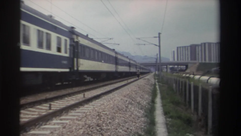 Cream-and-blue passenger coaches close to camera on electrified track, high-rise buildings and mountains behind, Taiwan, 1982.