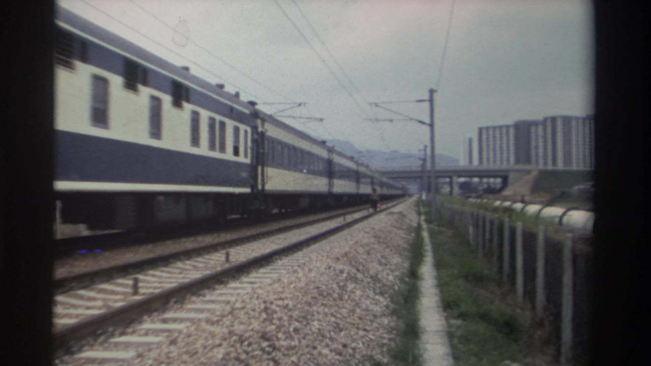 Blue-and-white passenger train on multi-track railway in what appears to be Taiwan, circa 1982, with concrete high-rises and mountain ridge beyond.