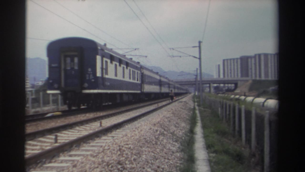 Rear carriage of blue passenger train moving away on multi-track railway, high-rise buildings and mountains in background.