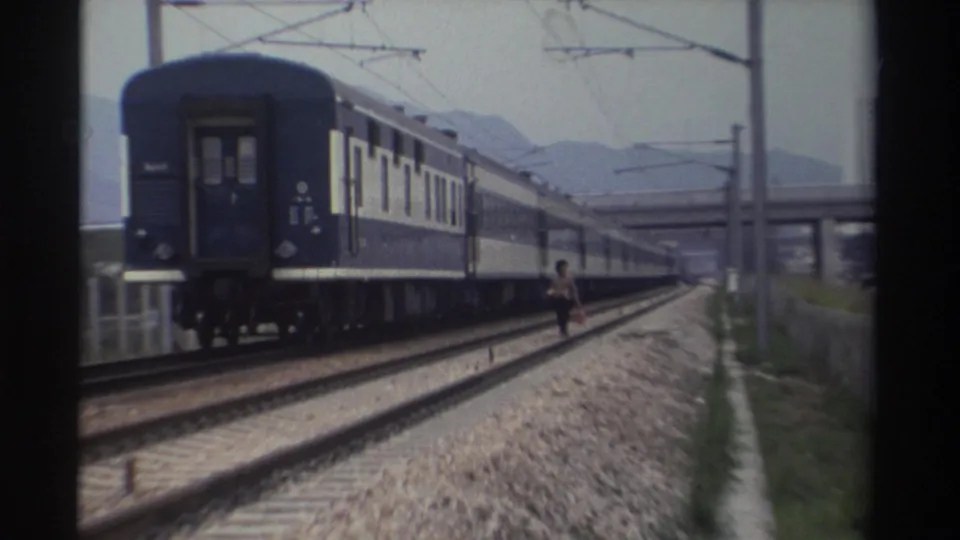 Adult figure walking beside a moving blue-and-white passenger train on what appears to be a Taiwanese railway, circa 1982, concrete high-rises beyond.