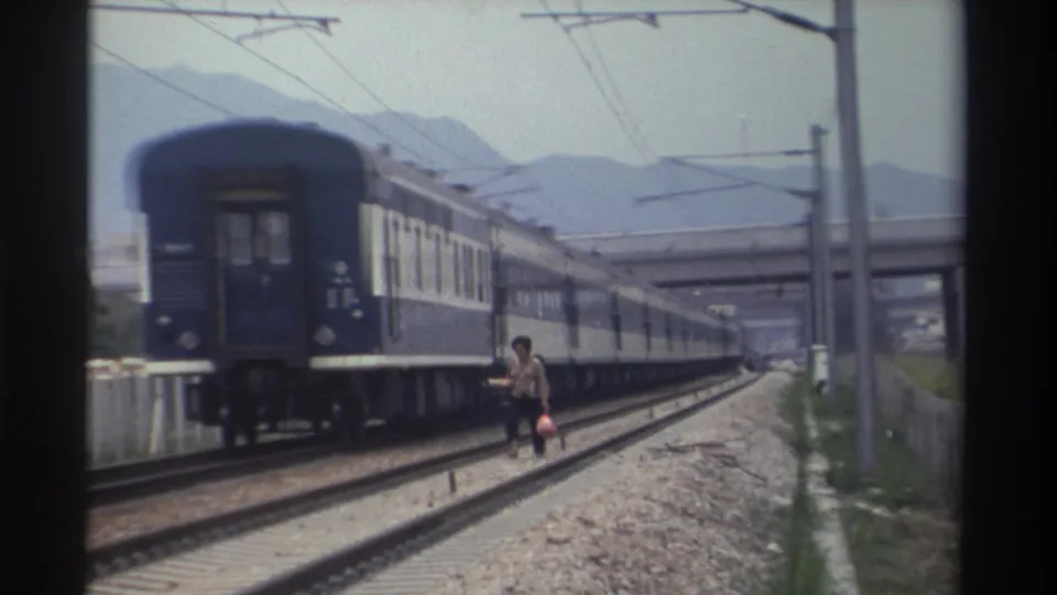 Adult figure with bag walking alongside a departing blue-and-white passenger train on what appears to be a Taiwanese railway, circa 1982.