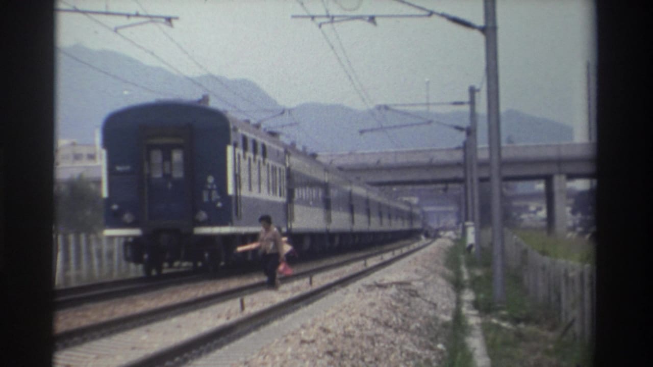 Adult figure with stick and bag beside an electrified railway line as a blue multi-car train passes; 1982 Taiwan, Super 8 home movie.