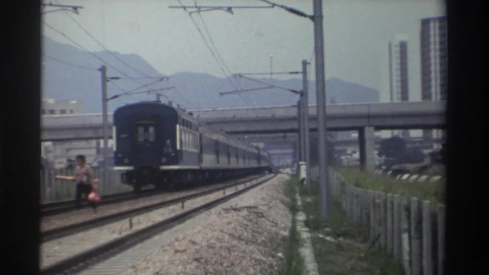 As the train clears frame, the trackside figure appears lower or crouching; mid-rise buildings and hazy mountain ridge visible; 1982 Taiwan.