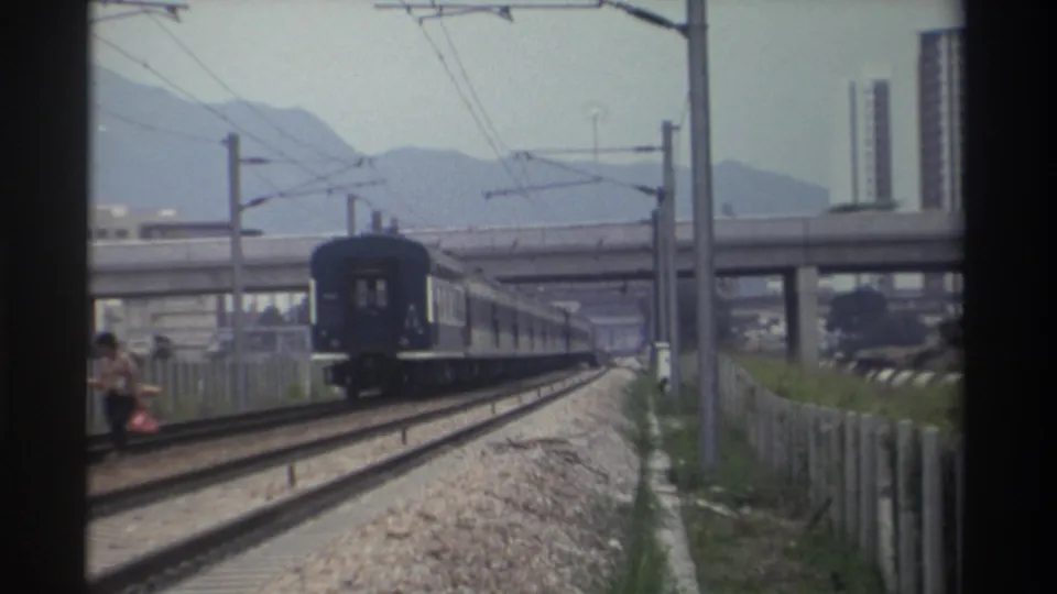 Closing frame as the last carriage exits; figure remains beside the tracks; mid-rise buildings and mountain skyline visible; 1982 Taiwan.