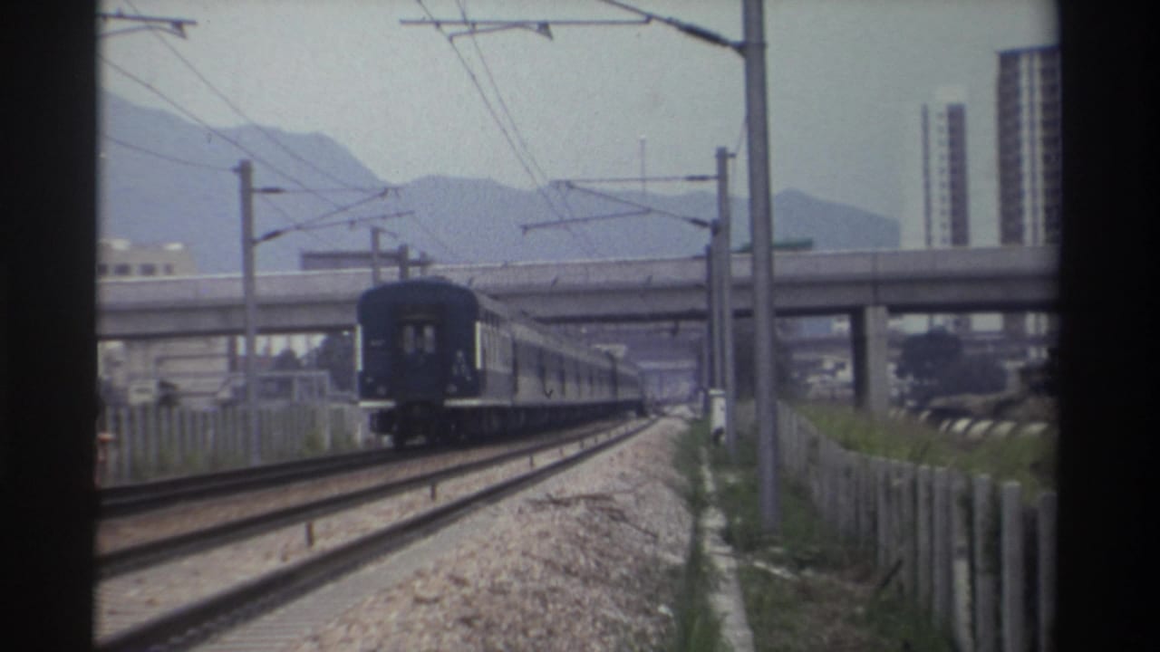 Dark blue passenger train moving along electrified railway in Taiwan, c.1982, concrete overpass mid-ground, mountain range and high-rise buildings behind.