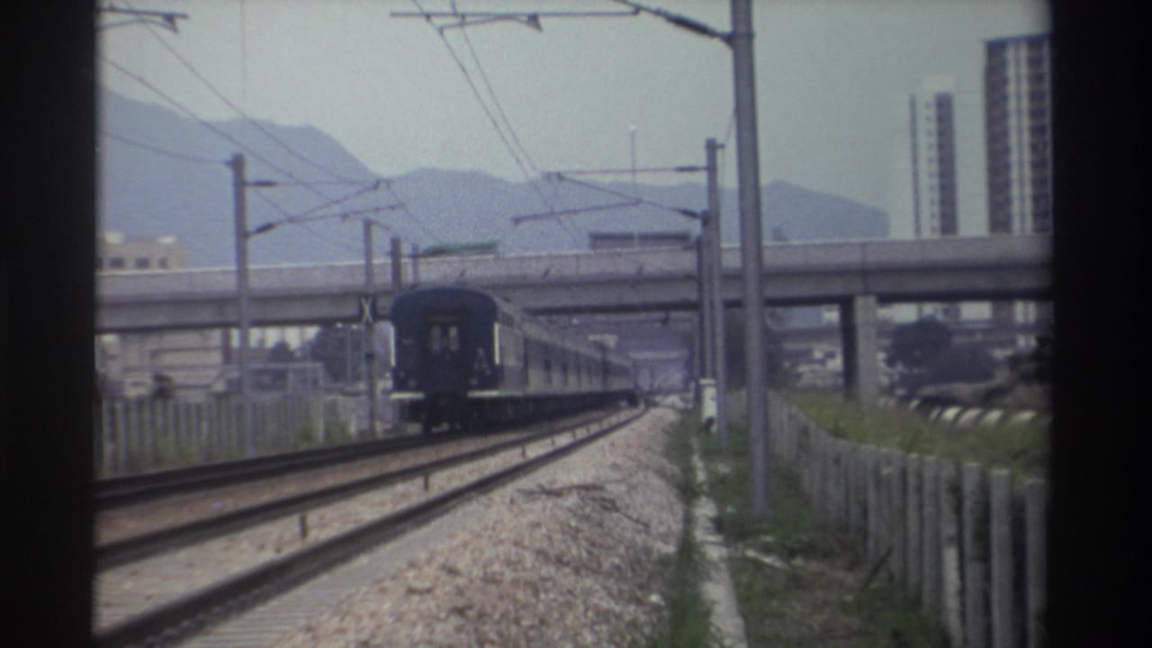 Blue passenger train on Taiwan railway, c.1982, advancing toward a concrete overpass with mountains and high-rise towers in the hazy background.