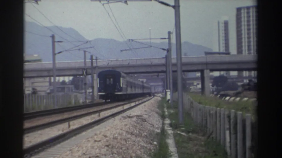 Blue passenger train passing under a concrete overpass on an electrified railway in Taiwan, c.1982; distant mountains and apartment blocks visible.