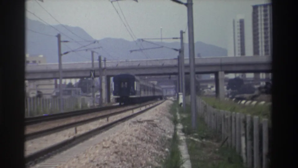 Rear carriages of blue passenger train receding along electrified railway in Taiwan, c.1982; concrete overpass, mountains and apartment towers remain in frame.