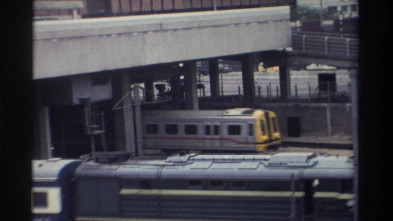 Silver commuter train with yellow end car and red accent stripe on elevated rails, urban Taiwan circa 1982, 8mm home-movie film.