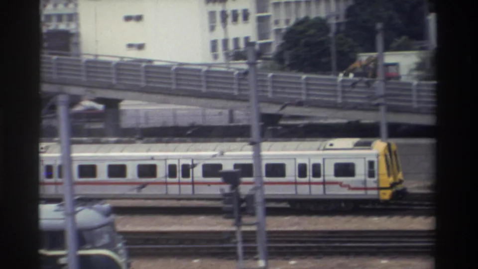Elevated rail yard with silver and yellow commuter train, mid-rise buildings and trees beyond, Taiwan circa 1982, 8mm film.