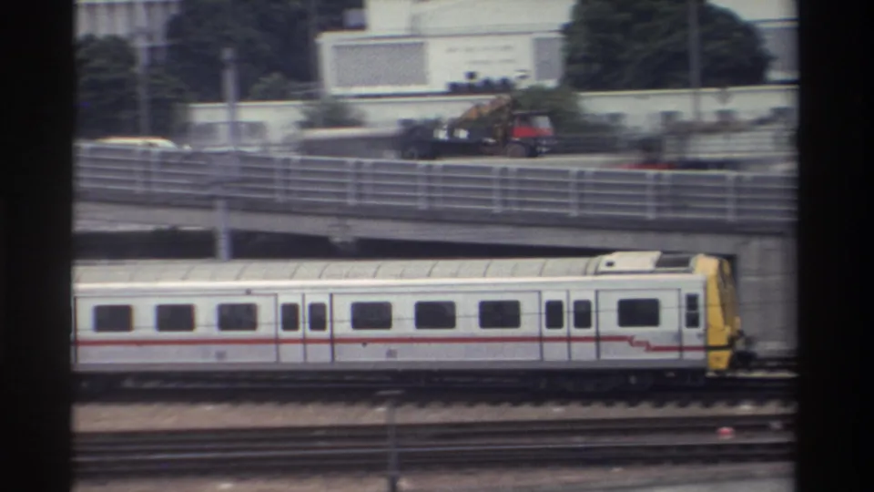 Silver and yellow commuter train on elevated rail infrastructure, mid-rise urban buildings behind, Taiwan circa 1982, 8mm home-movie film.