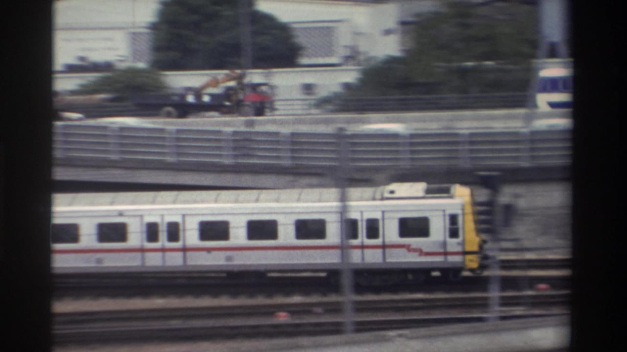 1982 Taiwan — white-and-yellow commuter train with red stripe passes beneath an overpass carrying a crane truck; 8mm film grain evident.