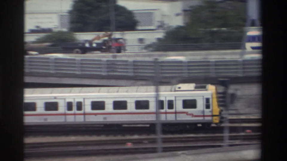 1982 Taiwan — white-and-yellow commuter train with red stripe passes beneath an overpass carrying a crane truck; 8mm film grain evident.
