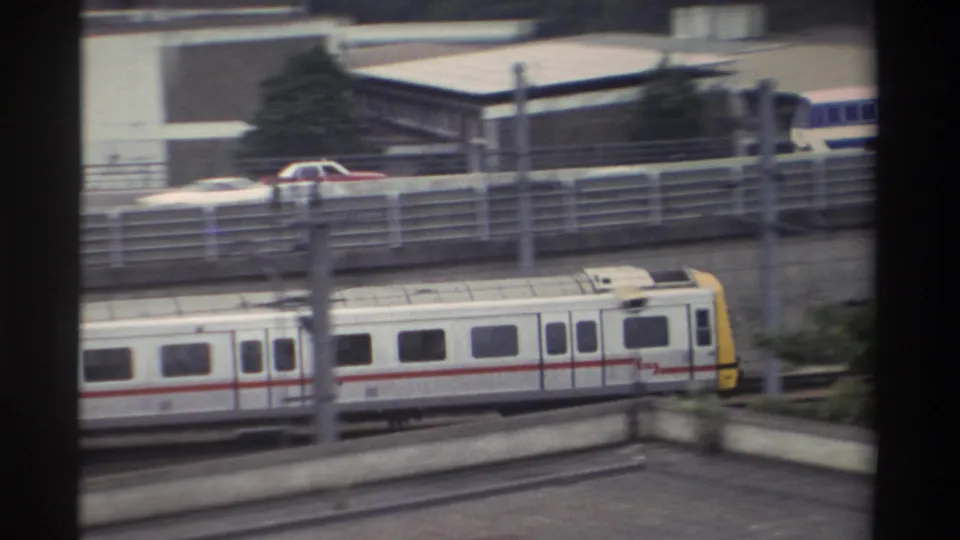 1982 Taiwan — train passing with vehicles including a red car on an adjacent road above a concrete retaining wall; urban setting.