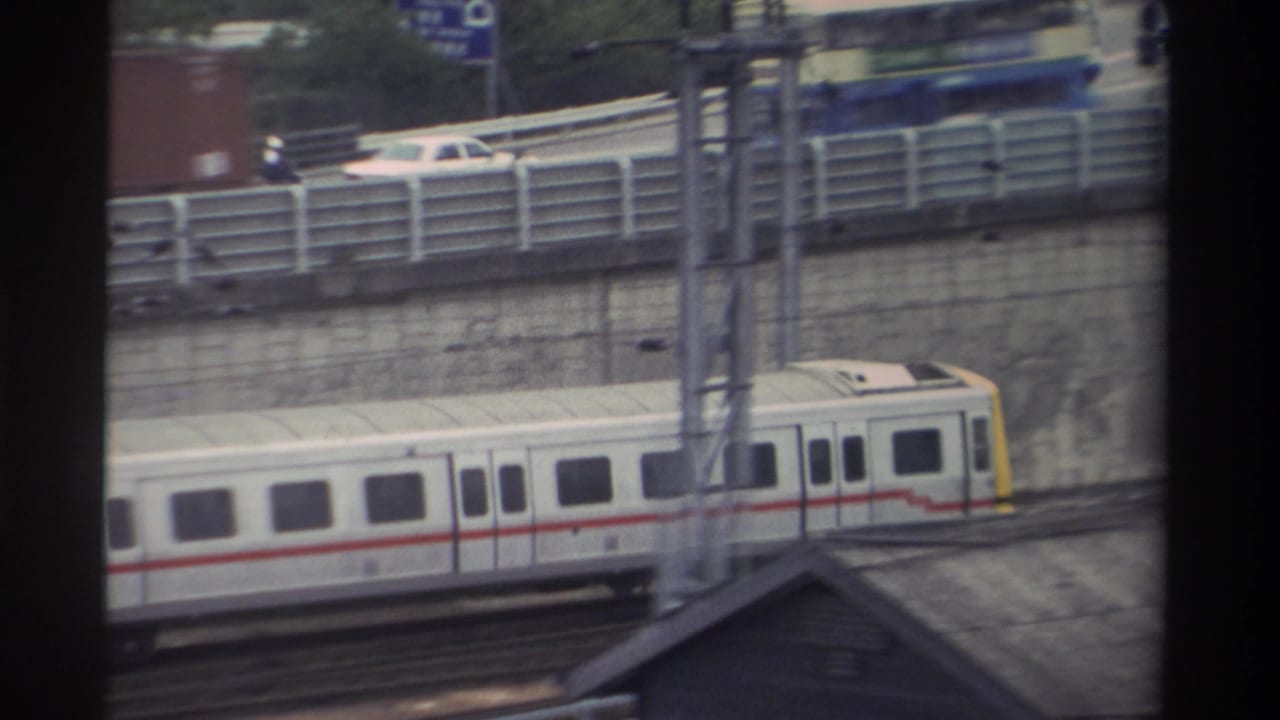 White passenger train with red stripe and yellow cab on elevated track in Taiwan, 1982; highway traffic and a partially visible blue road sign below, shot through window mullions.