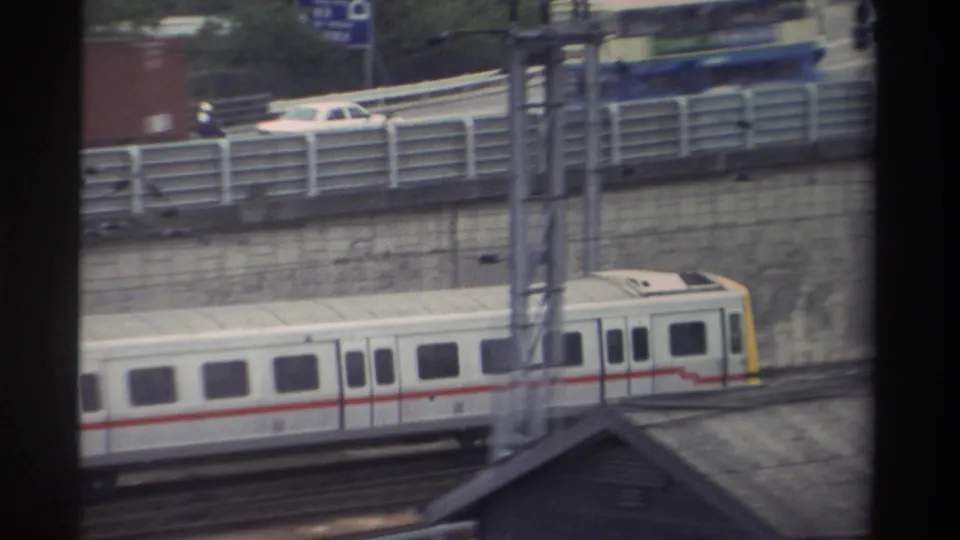 White passenger train with red stripe and yellow cab on elevated track in Taiwan, 1982; highway traffic and a partially visible blue road sign below, shot through window mullions.