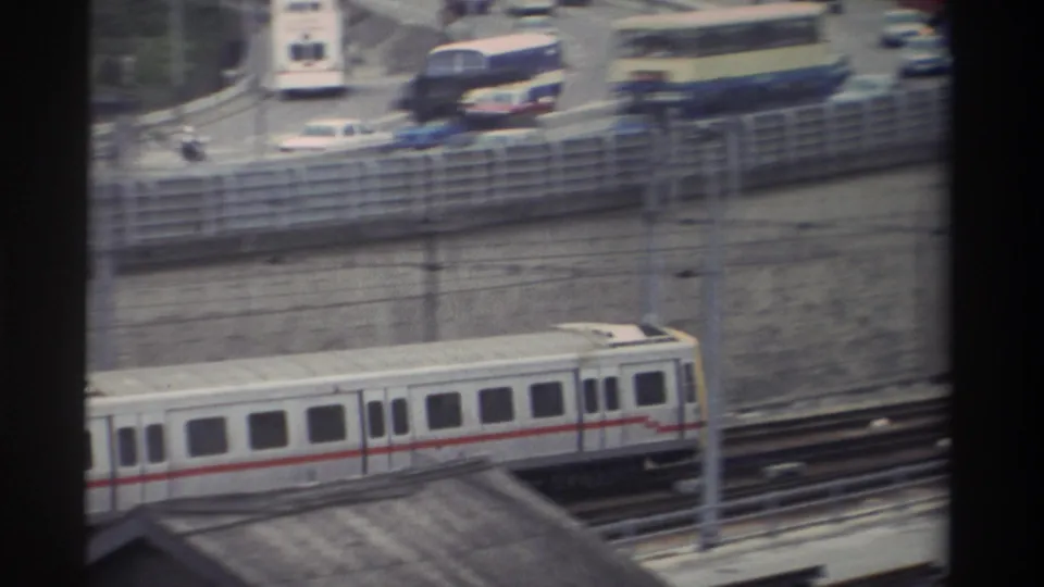 White passenger train with red stripe alongside busy highway with colorful buses and mixed traffic, Taiwan, 1982, framed through window.