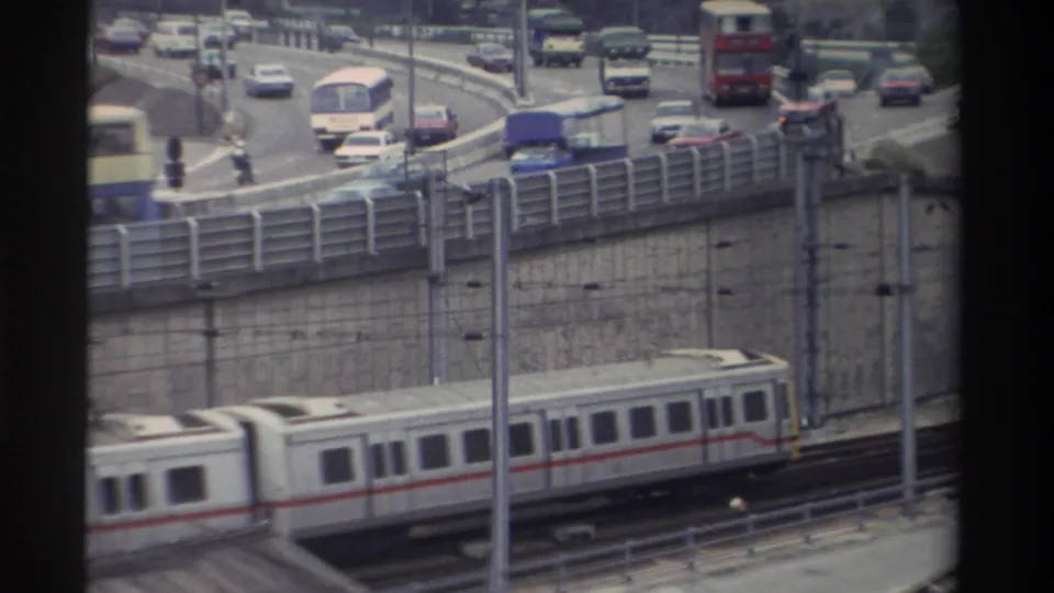 White passenger train with red stripe beside busy multi-lane highway, Taiwan, 1982; mixed vehicle traffic and catenary poles visible through window frame.