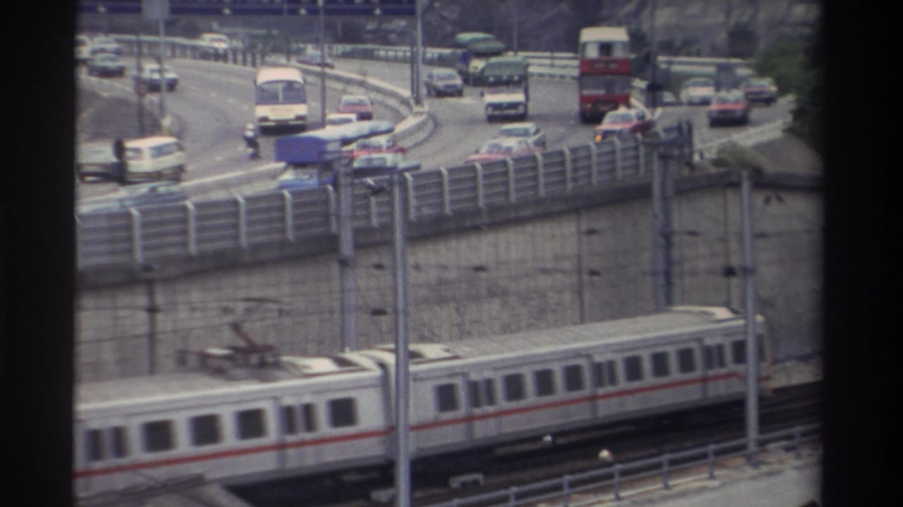 8mm home movie frame: white and red-striped light rail train beneath a curved elevated highway carrying a red double-decker bus and saloon cars, Taiwan, 1982.
