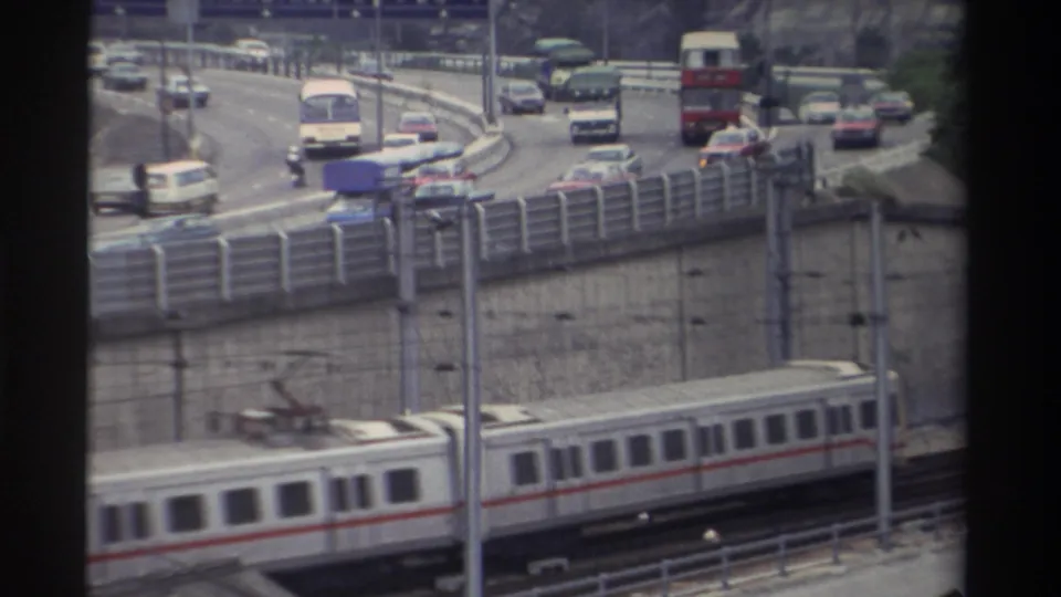 8mm home movie frame: white and red-striped light rail train beneath a curved elevated highway carrying a red double-decker bus and saloon cars, Taiwan, 1982.