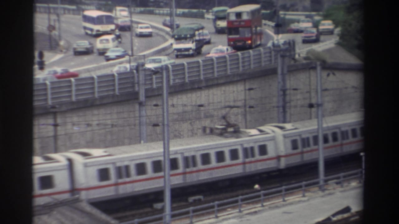 8mm footage: white and red-striped light rail cars below a multi-lane highway ramp with red double-decker bus, saloon cars, and a white van, Taiwan, 1982.