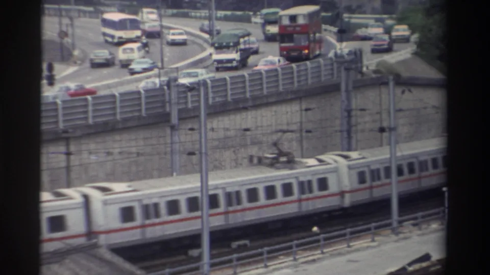 8mm footage: white and red-striped light rail cars below a multi-lane highway ramp with red double-decker bus, saloon cars, and a white van, Taiwan, 1982.