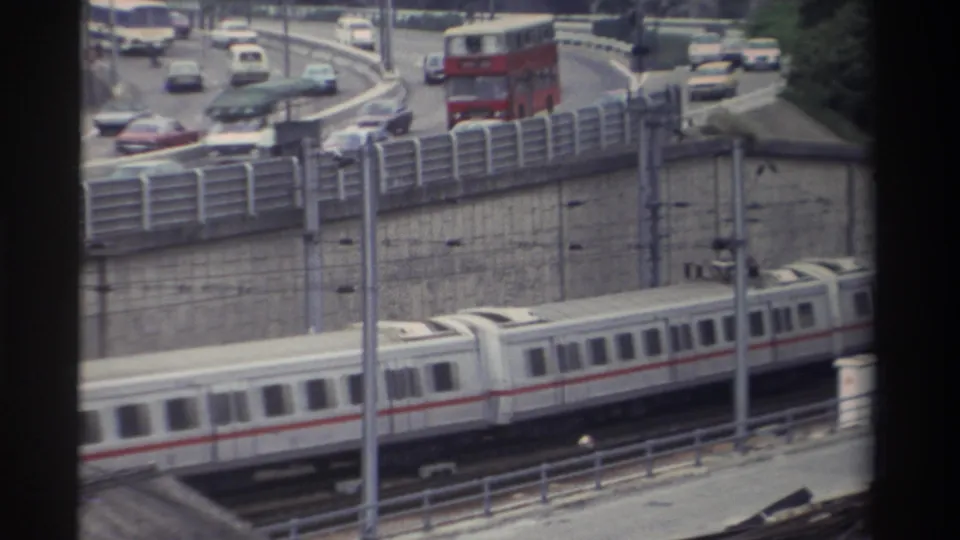 8mm home movie: red double-decker bus on a curved elevated highway ramp, light rail train below, yellow car visible among mixed saloon traffic, Taiwan, 1982.
