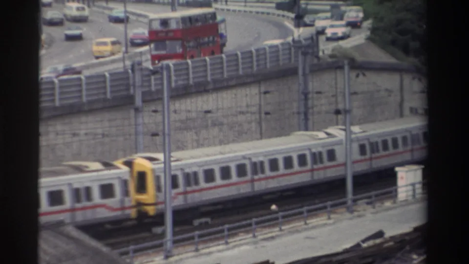 8mm frame: yellow-fronted light rail cab moving beneath a multi-lane elevated highway with a red double-decker bus and yellow vehicle among saloon car traffic, Taiwan, 1982.
