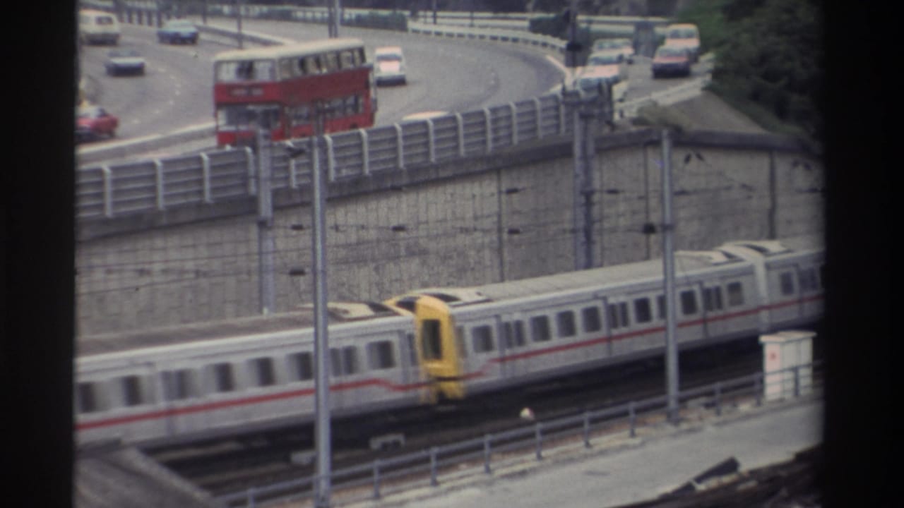 White and red passenger train with yellow front cab moving along tracks; red double-decker bus visible on curved concrete road above.