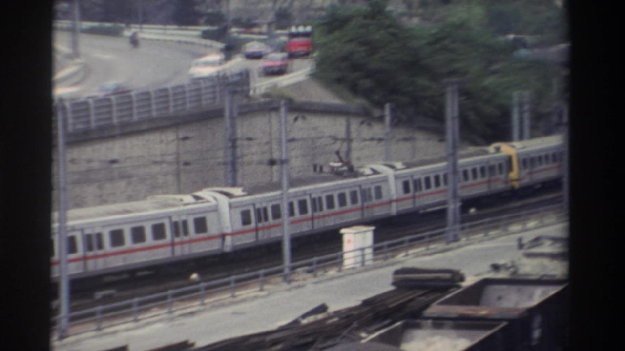 1982 Super 8 home movie from Taiwan: long passenger train with red horizontal stripe and yellow front car passing on elevated tracks beside a curved road.