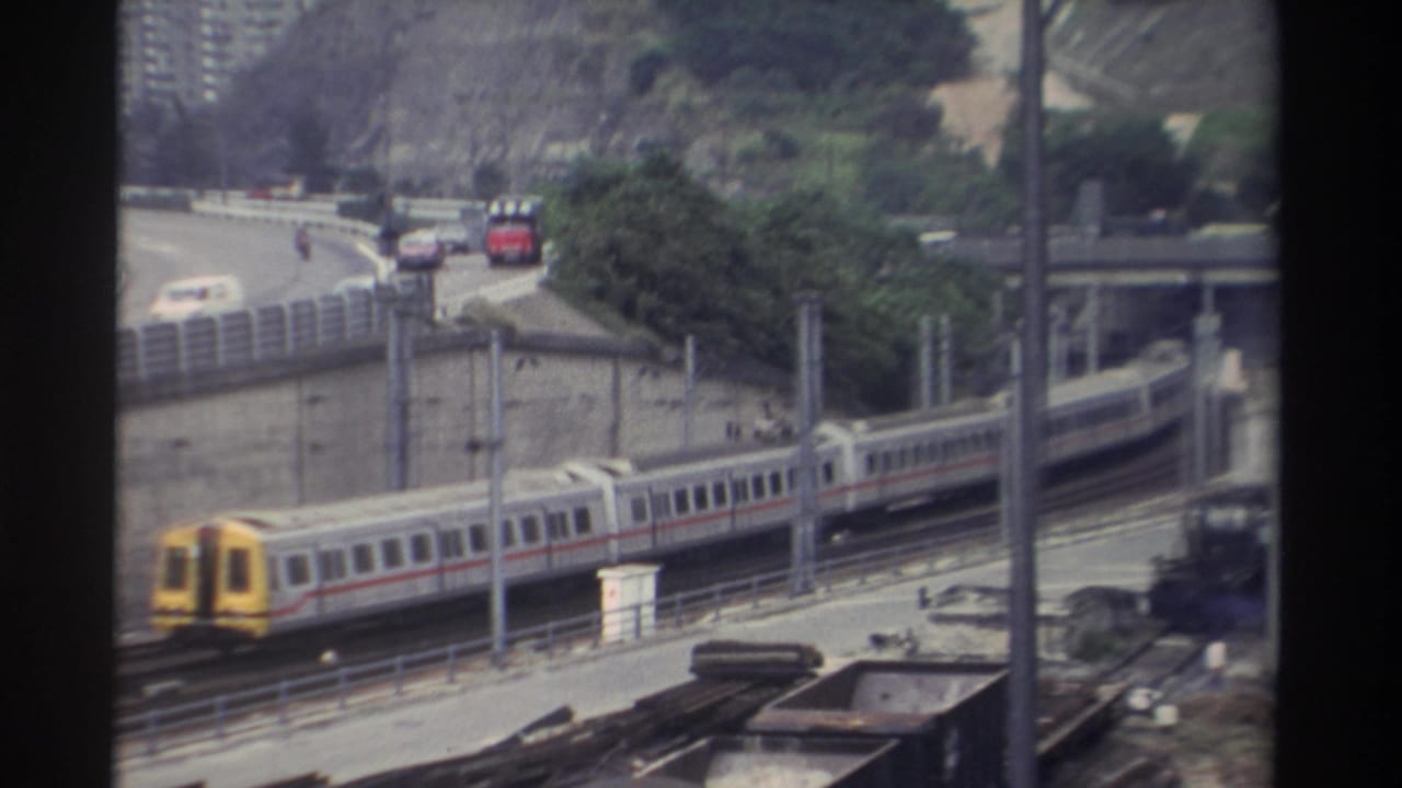 Super 8 footage circa 1982, Taiwan: train with yellow cab moving along railway corridor with highway traffic and tree-covered hillside behind.