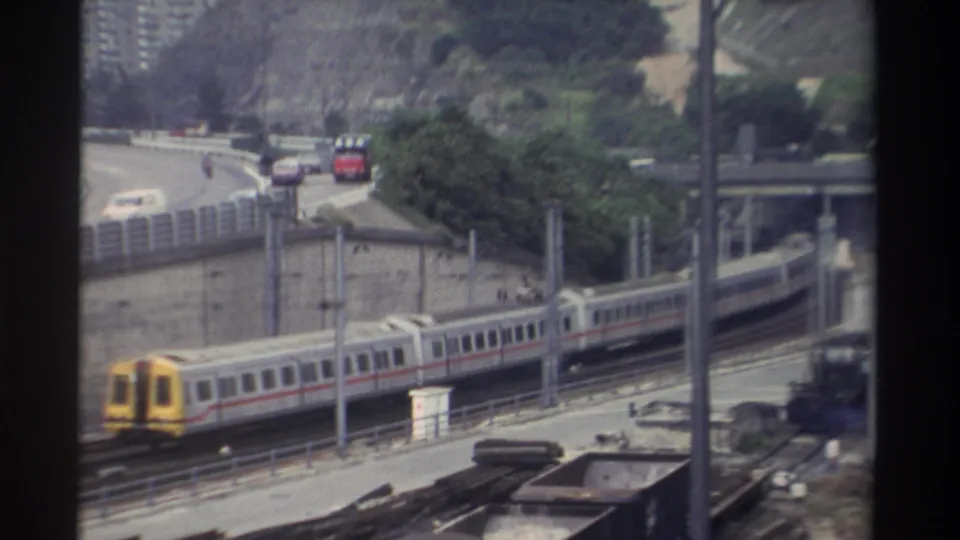 Super 8 footage circa 1982, Taiwan: train with yellow cab moving along railway corridor with highway traffic and tree-covered hillside behind.