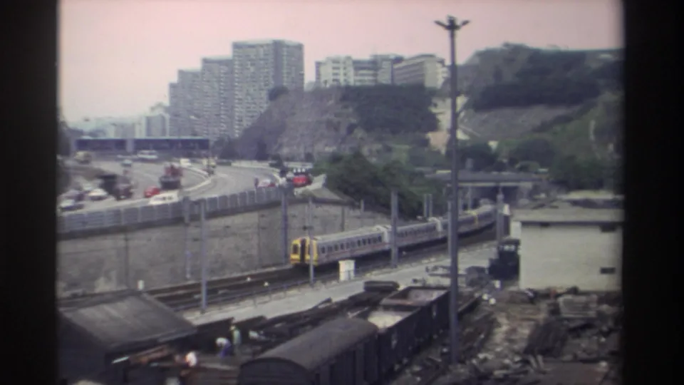1982 Super 8 home movie, Taiwan: wide urban scene with passenger train, highway traffic, concrete infrastructure, and high-rise housing on a hillside; faded pink film color.